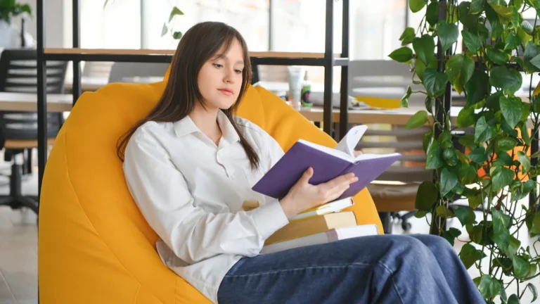 Jeune femme lisant un livre installée dans un grand pouf jaune, près de plantes vertes